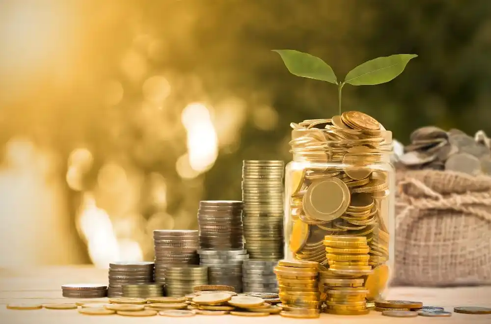 A jar filled with money and coins sits on a wooden table, with a small plant growing from the top, symbolizing growth and savings.